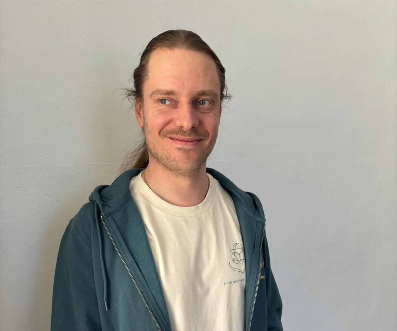 Ville Vertanen seisoo valkoisen seinän edessä ja katsoo hymyillen sivulle. 